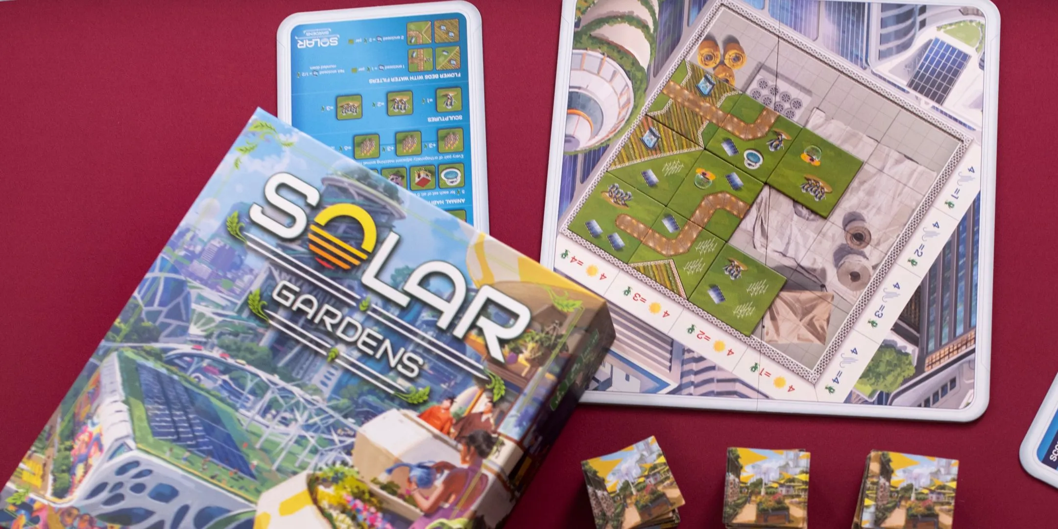 Các thành phần game Solar Gardens bao gồm hộp, bảng người chơi và ô gạch được sắp đặt trên nền đỏ, thể hiện sự sang trọng và thân thiện với môi trường